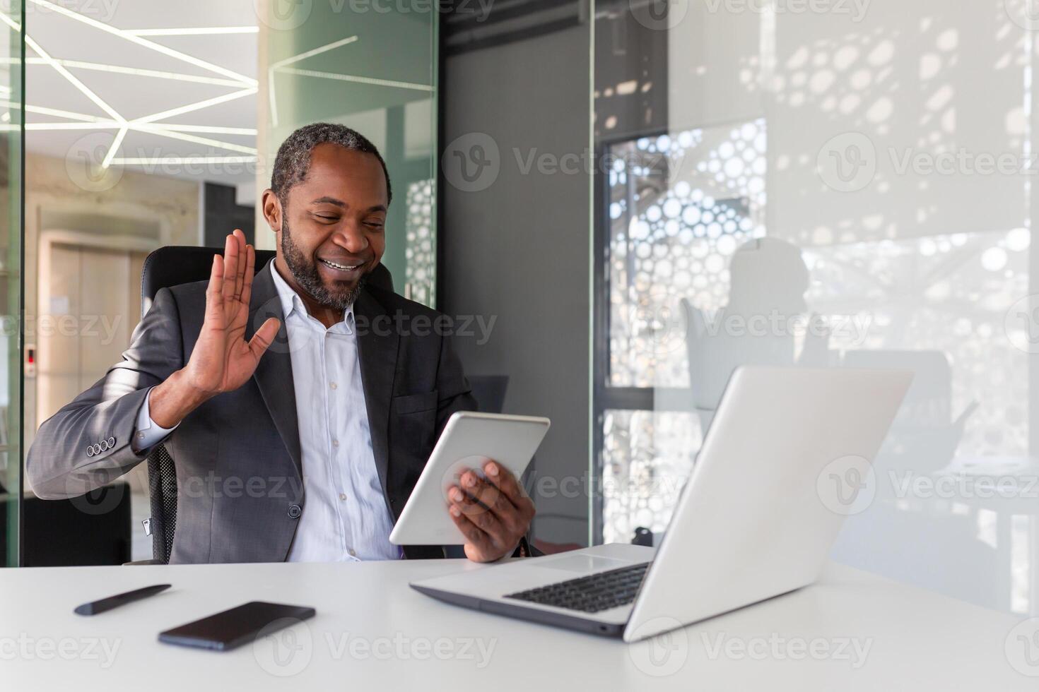 Mature successful boss inside office at workplace using tablet computer for call, man sitting at table with laptop, waving joyfully at device camera, using app for remote communication. photo