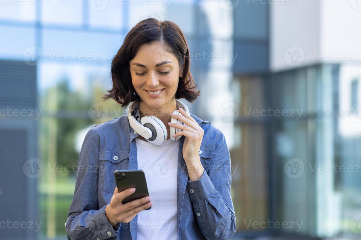 A young female student with a cheerful expression uses her smartphone and wears headphones, standing outdoors near a modern building. Concept of technology in daily life. photo