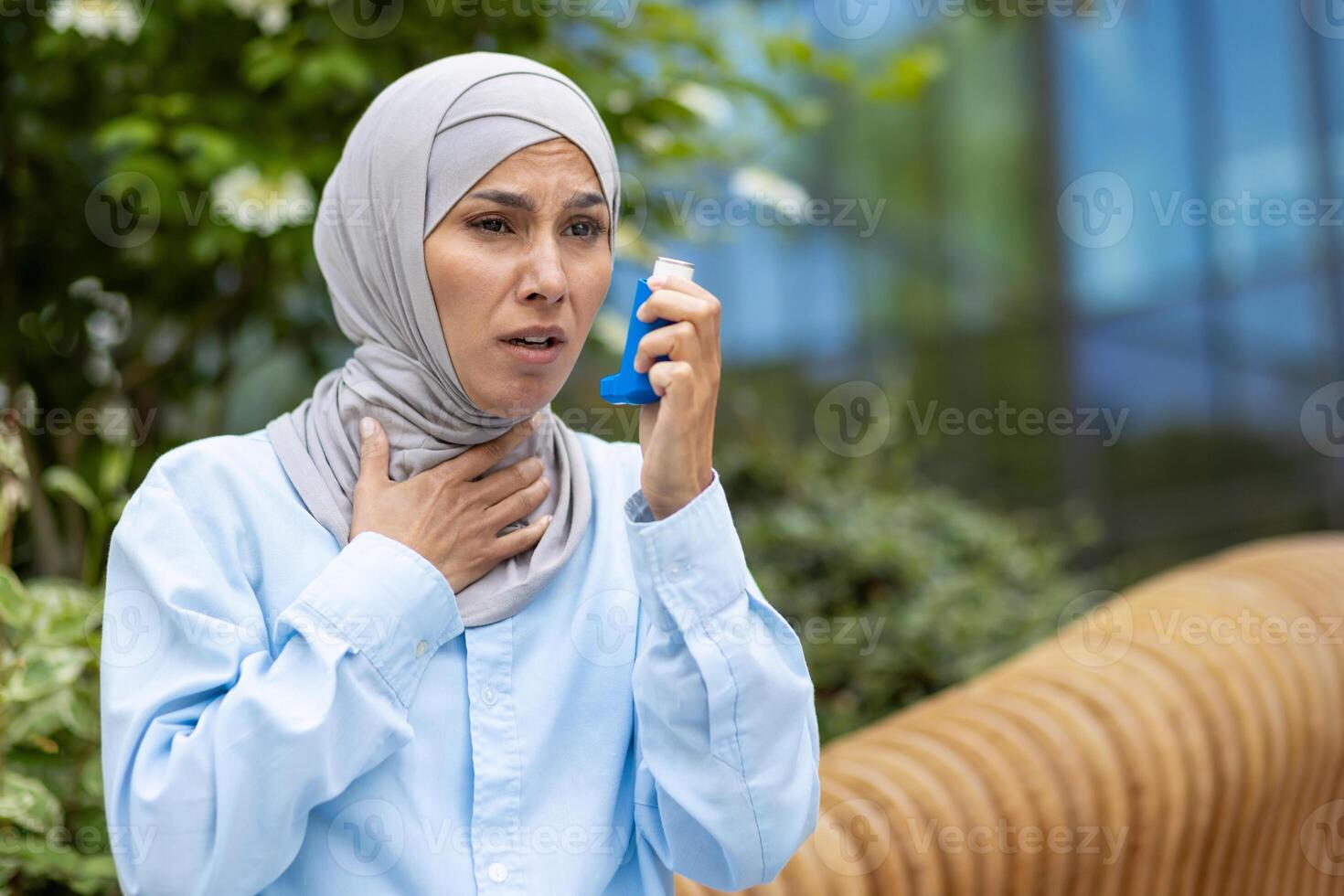 Shocked arabic woman covering neck with hand and holding bronchial asthma inhaler while having walk outdoors. Scared lady suffering from seasonal allergy and feeling hard to breath without medicine. photo