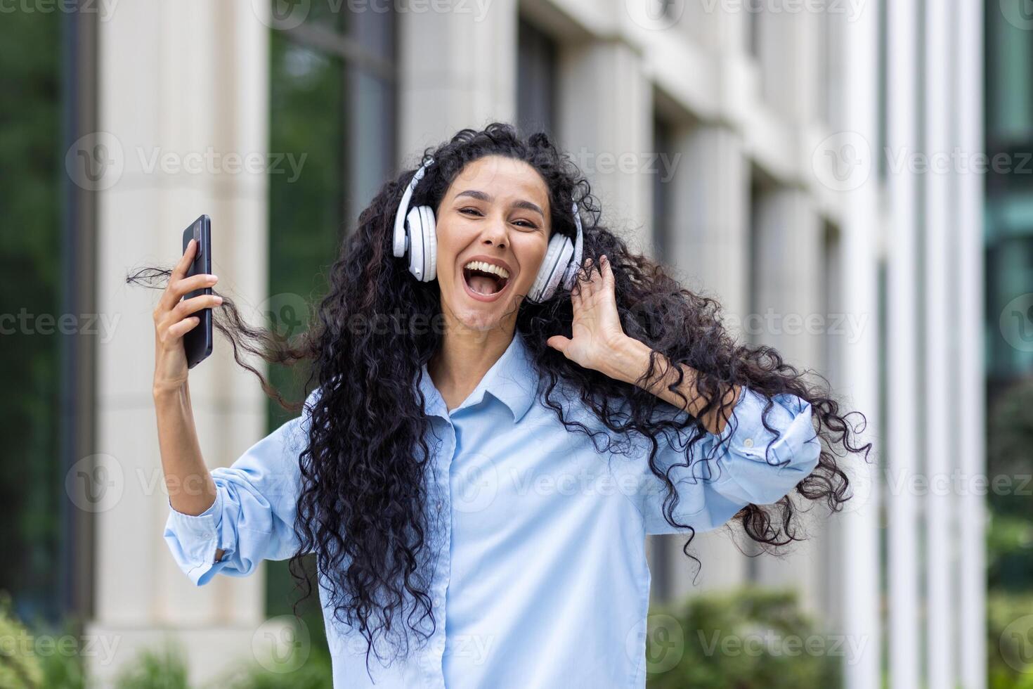 An energetic young woman with long curly hair, enjoying her favorite tunes with headphones, holding a smartphone outdoors with office buildings in the background, conveying a sense freedom and joy. photo