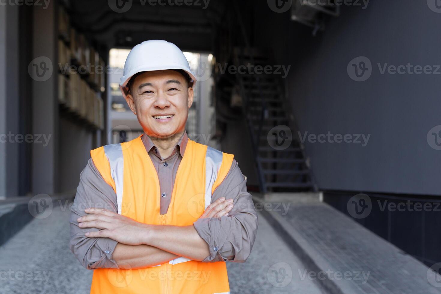 Close-up portrait of an Asian young man in a construction helmet and vest, an employee of an engineering firm, an architect standing in front of the camera smiling and crossing his arms over his chest. photo