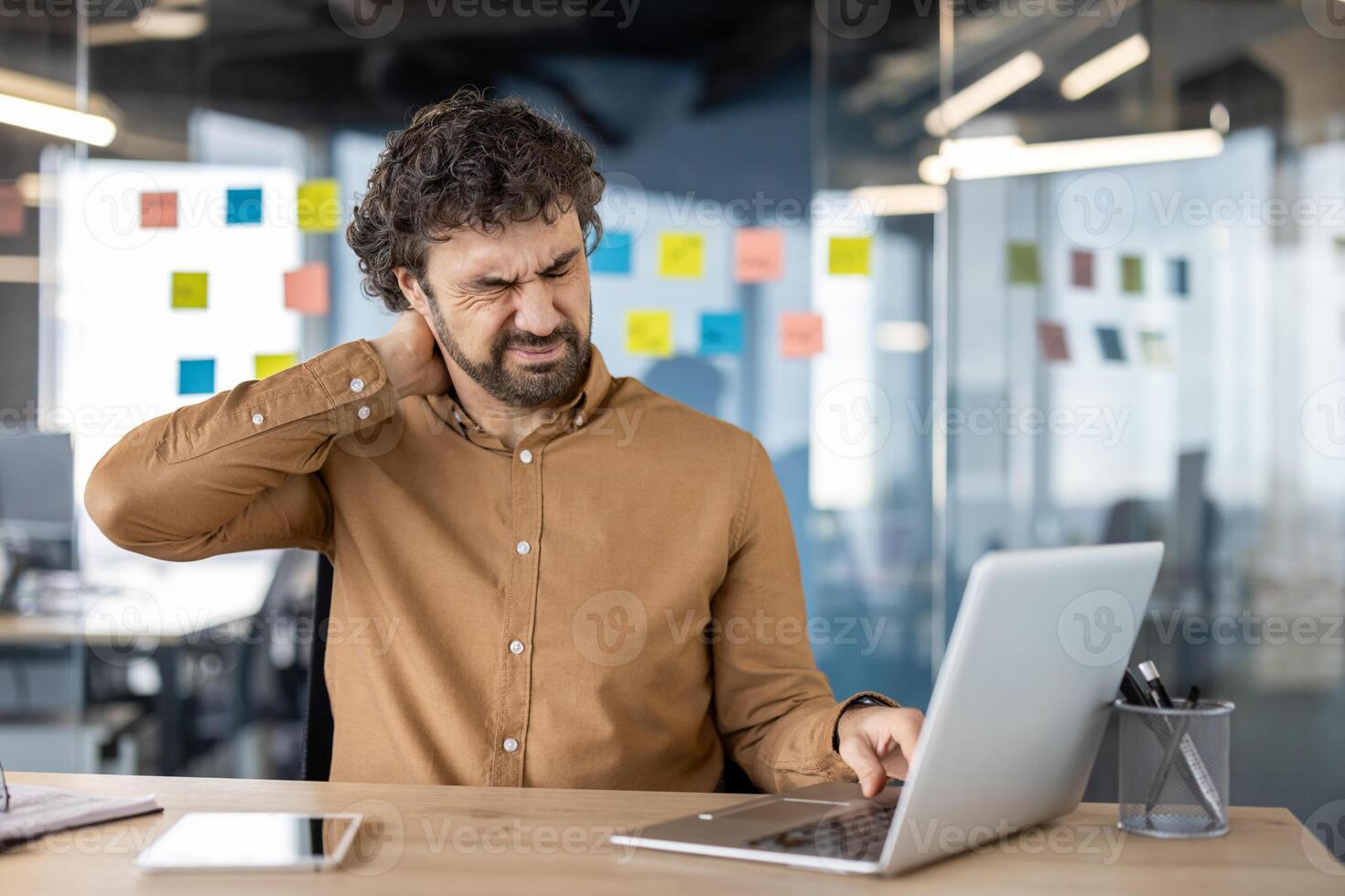 A male office employee feels sharp neck pain while working on his laptop, indicating the need for ergonomic workspaces. photo