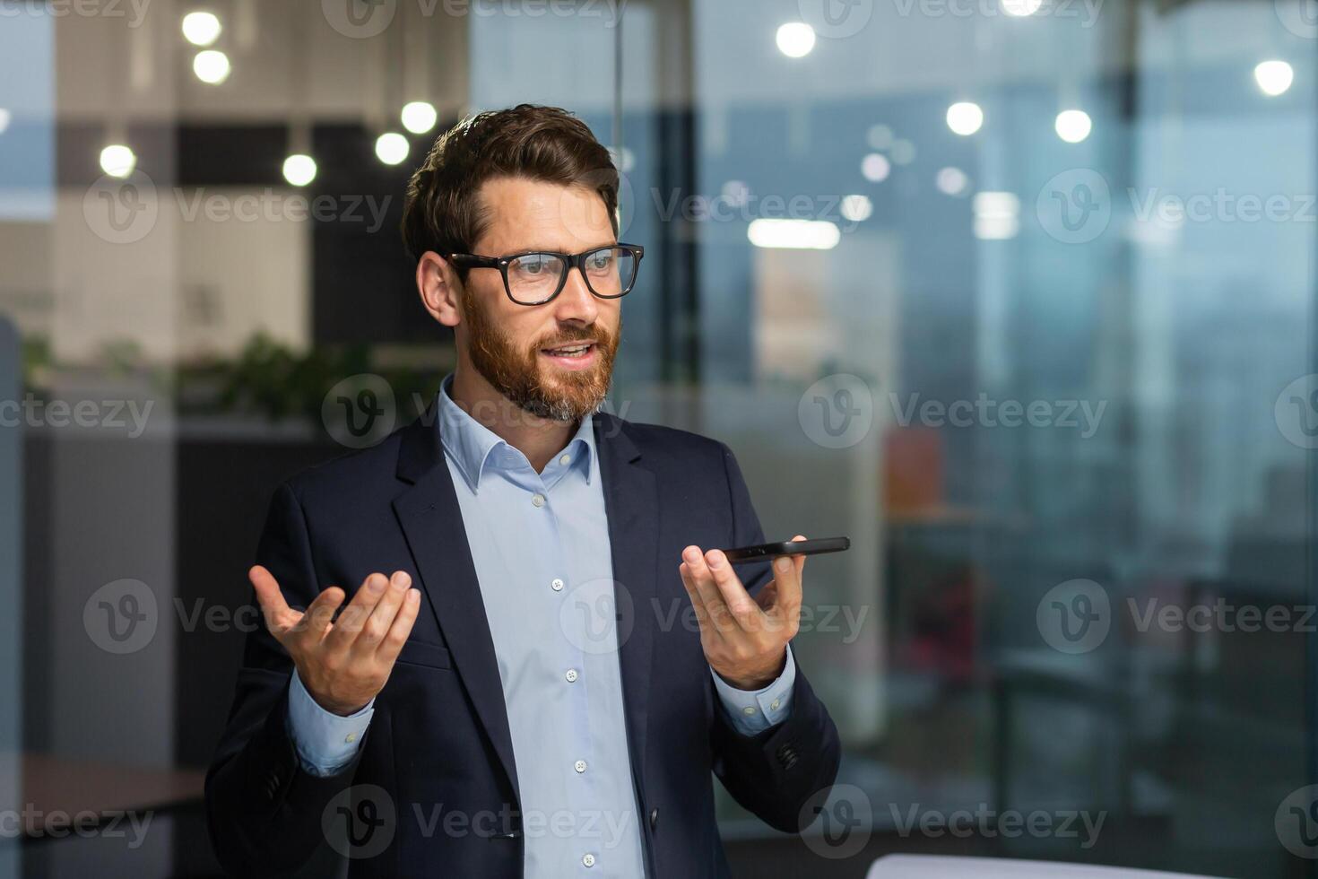 Friendly and smiling businessman in office recording audio message, man using smartphone app for communication, investor in business suit near window inside building. photo