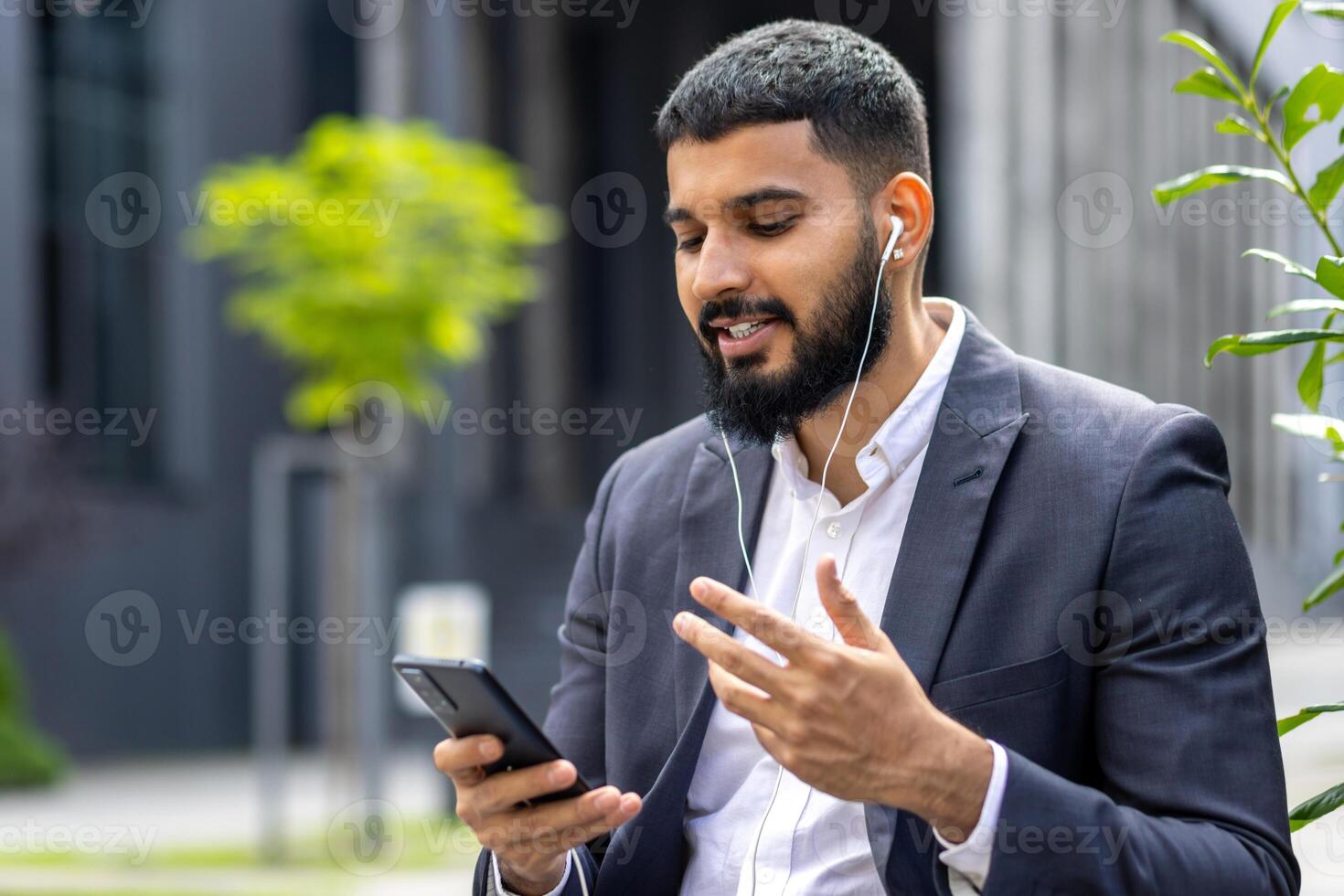 A well-dressed man engages with his smartphone while using earphones outside a modern building, possibly during a work break or in transit. photo