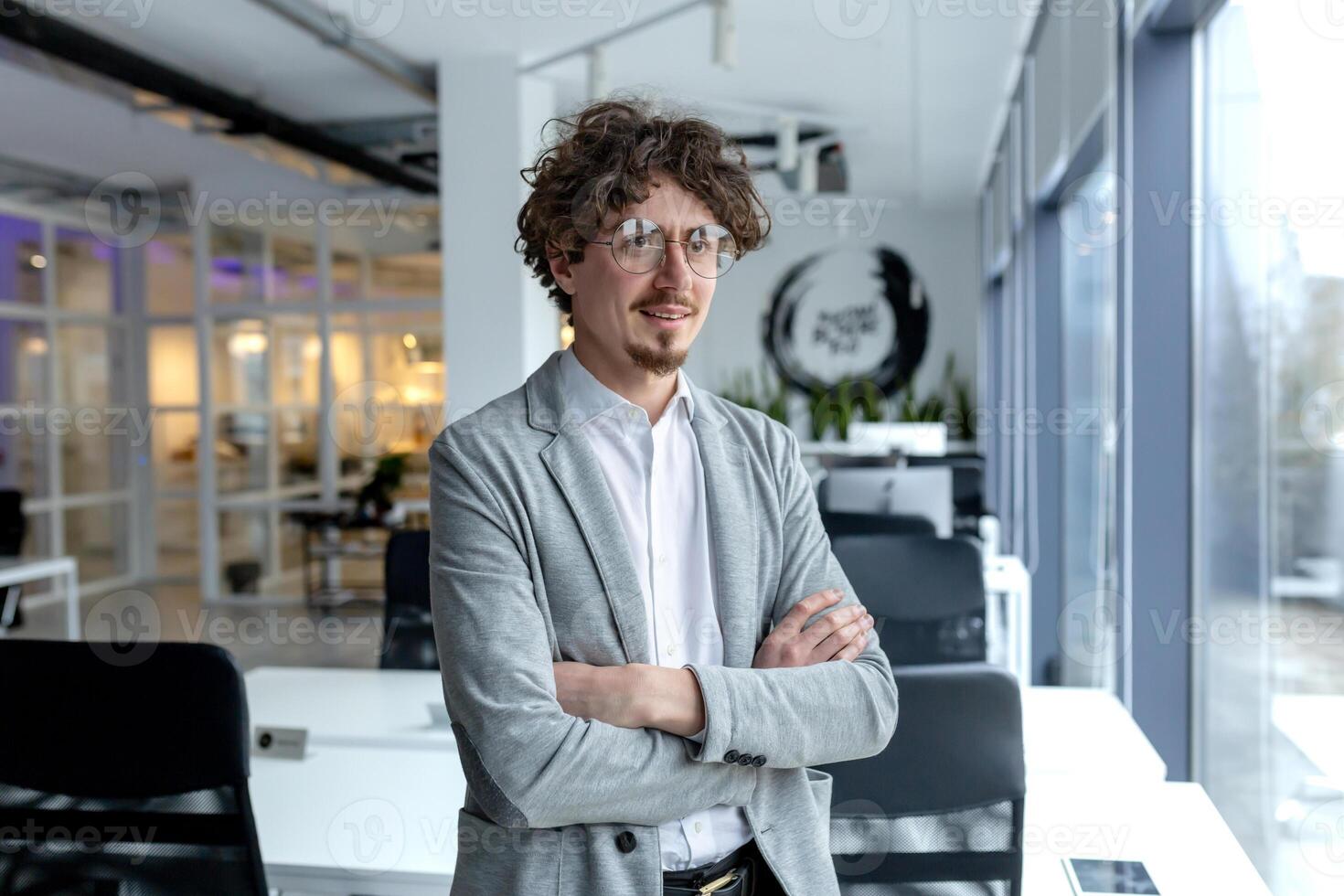 Professional young male entrepreneur with curly hair stands confidently in a well-lit office space, displaying experience and competence. photo