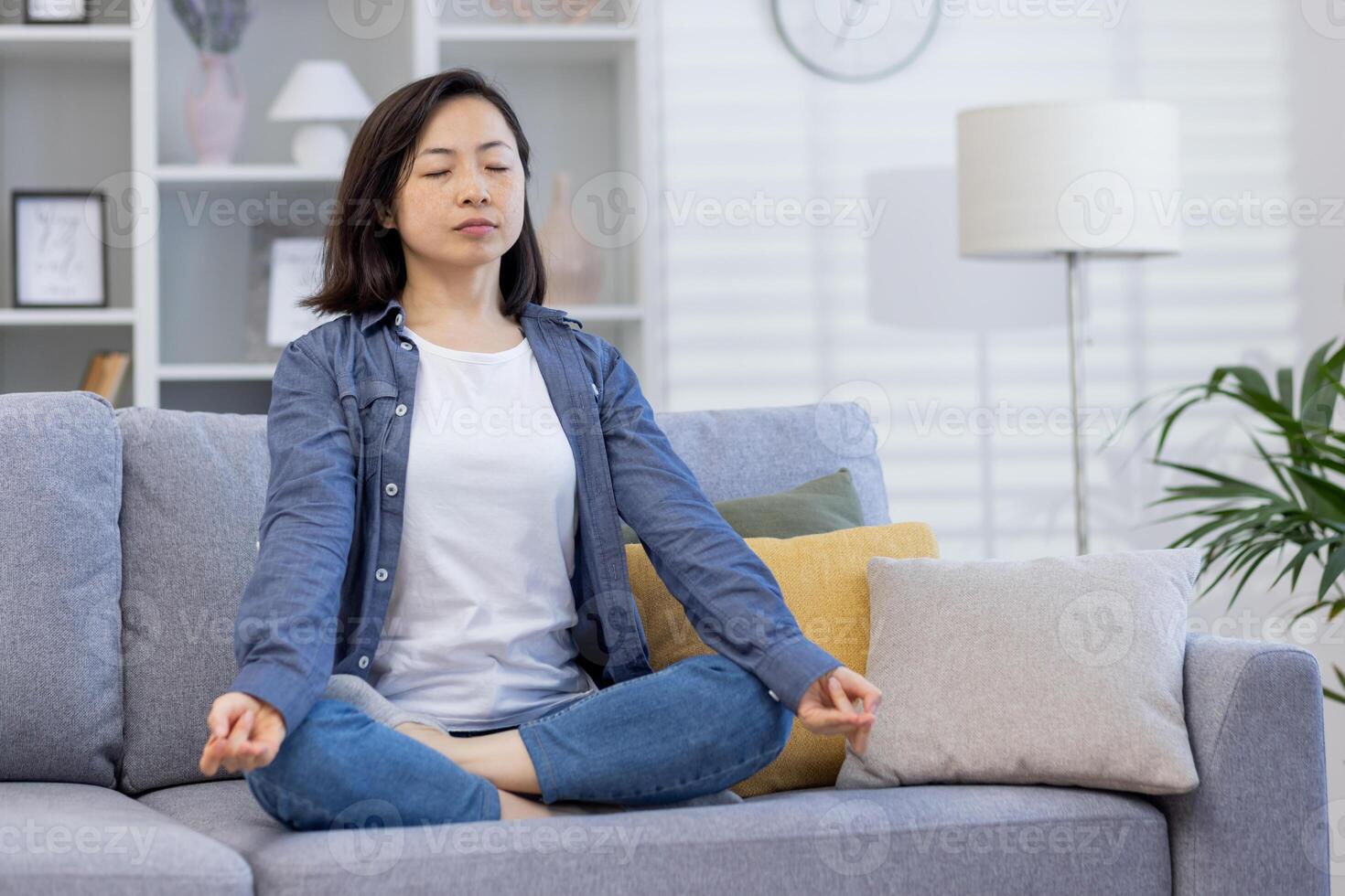 joven hermosa asiático mujer solo a hogar, meditando sentado en sofá en vivo habitación en loto posición, sentado con ojos cerrado a hogar. foto