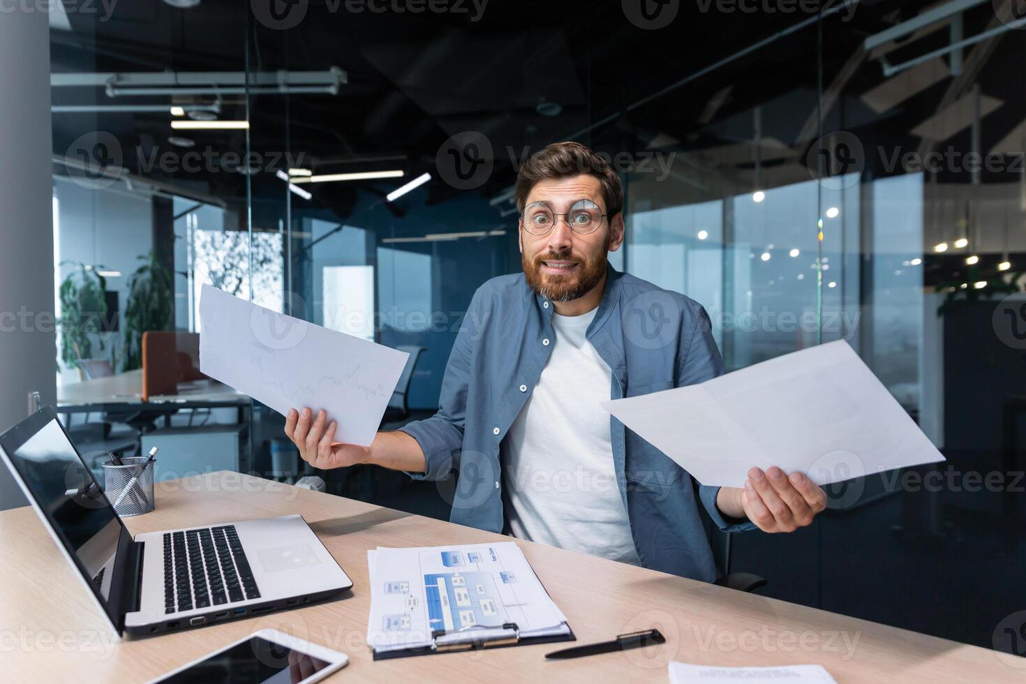 Dissatisfied and angry businessman inside office showing reports and bills documents to camera, boss in shirt looking at camera working with laptop paper work. photo