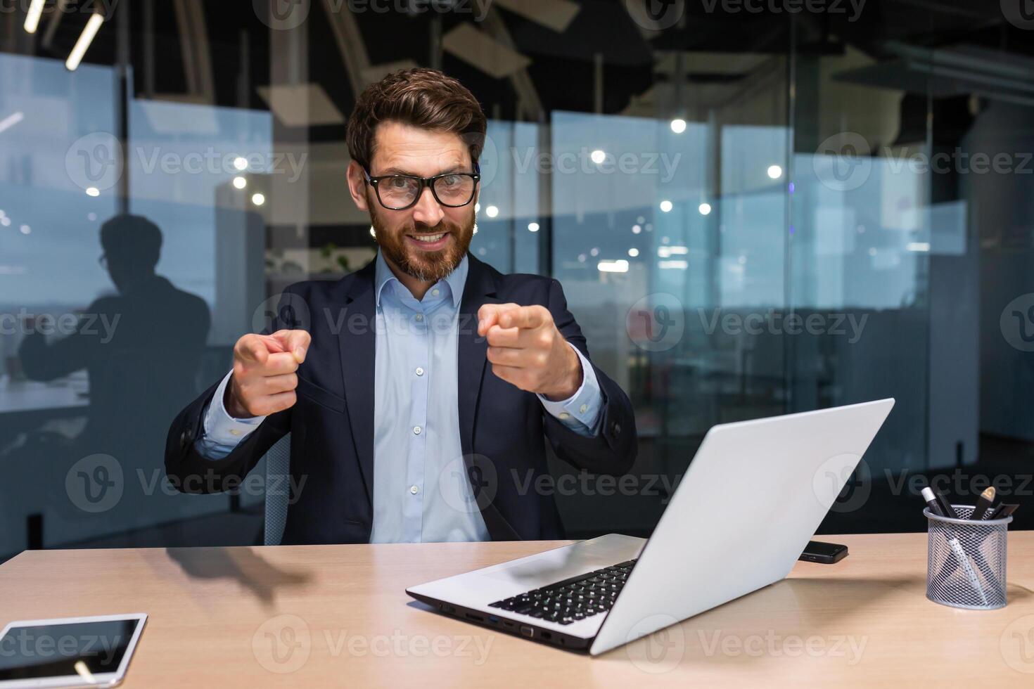 Successful mature businessman looking at camera and pointing finger forward calling to action, investor in business suit inside modern office working on laptop, boss with beard and glasses. photo