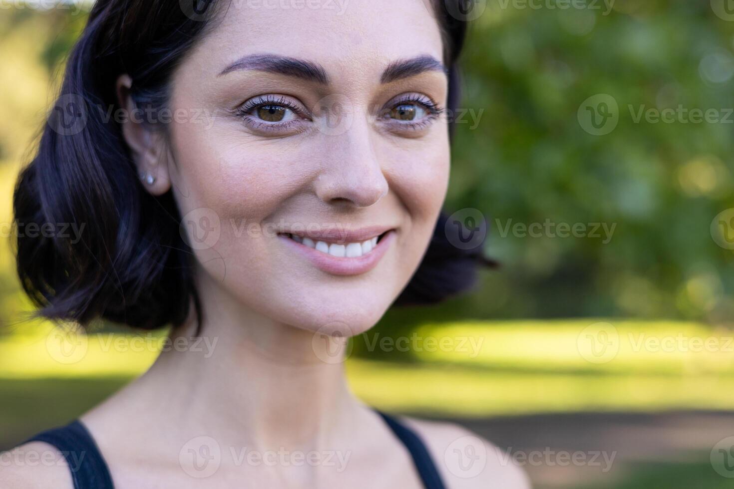 Close-up of a cheerful young woman smiling in nature with sunlight filtering through the trees. photo