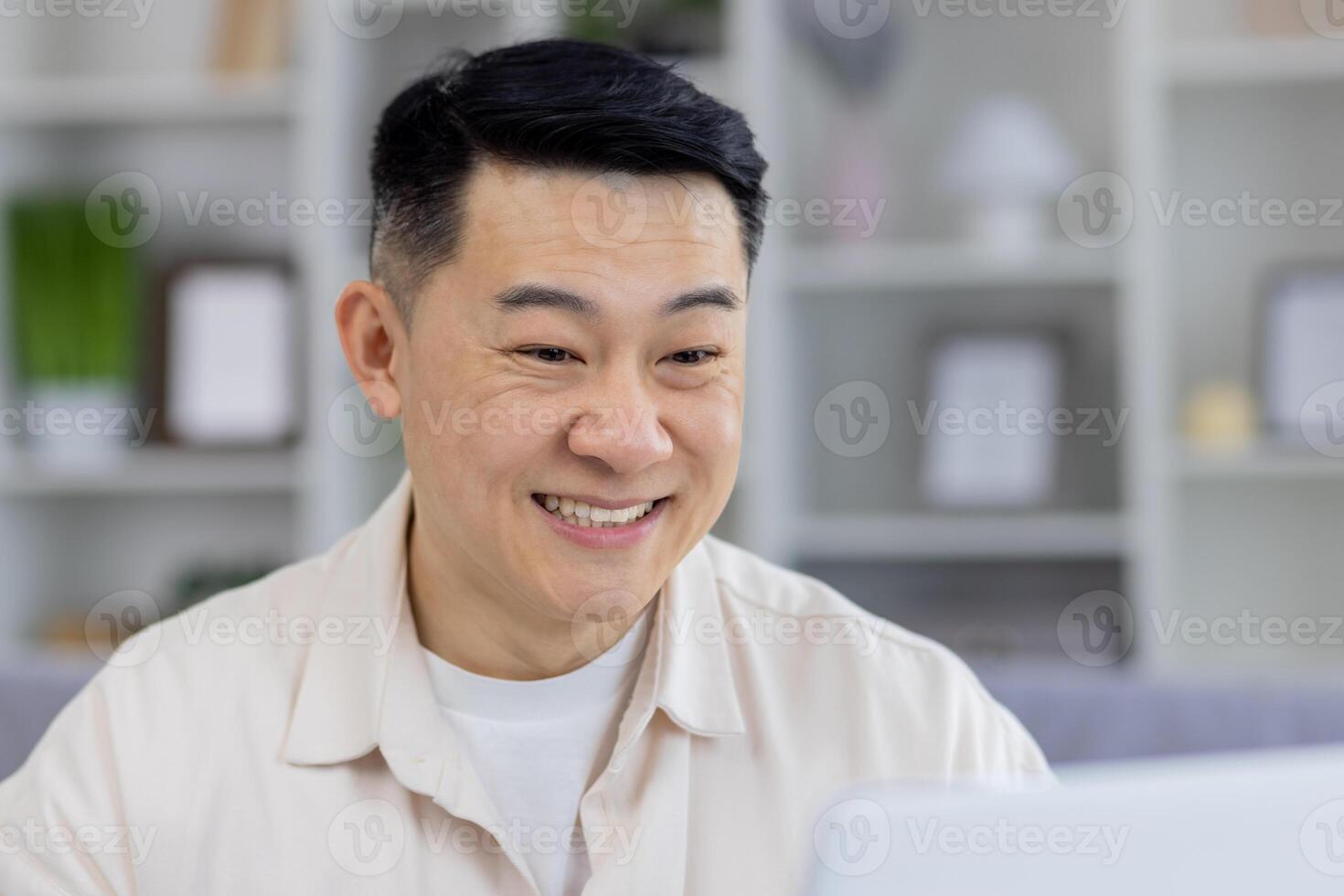 A middle-aged Asian man showing a joyful expression while interacting on his laptop in a stylishly decorated living room. Perfect imagery for themes on remote work, happiness, and technology use at home. photo