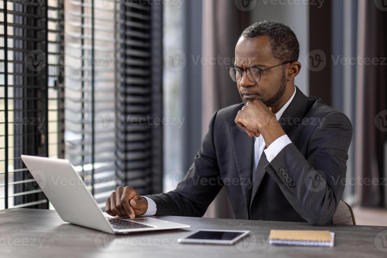 A professional businessman in a suit concentrating on his work on a laptop at a modern office desk, possibly analyzing data or planning strategies. photo
