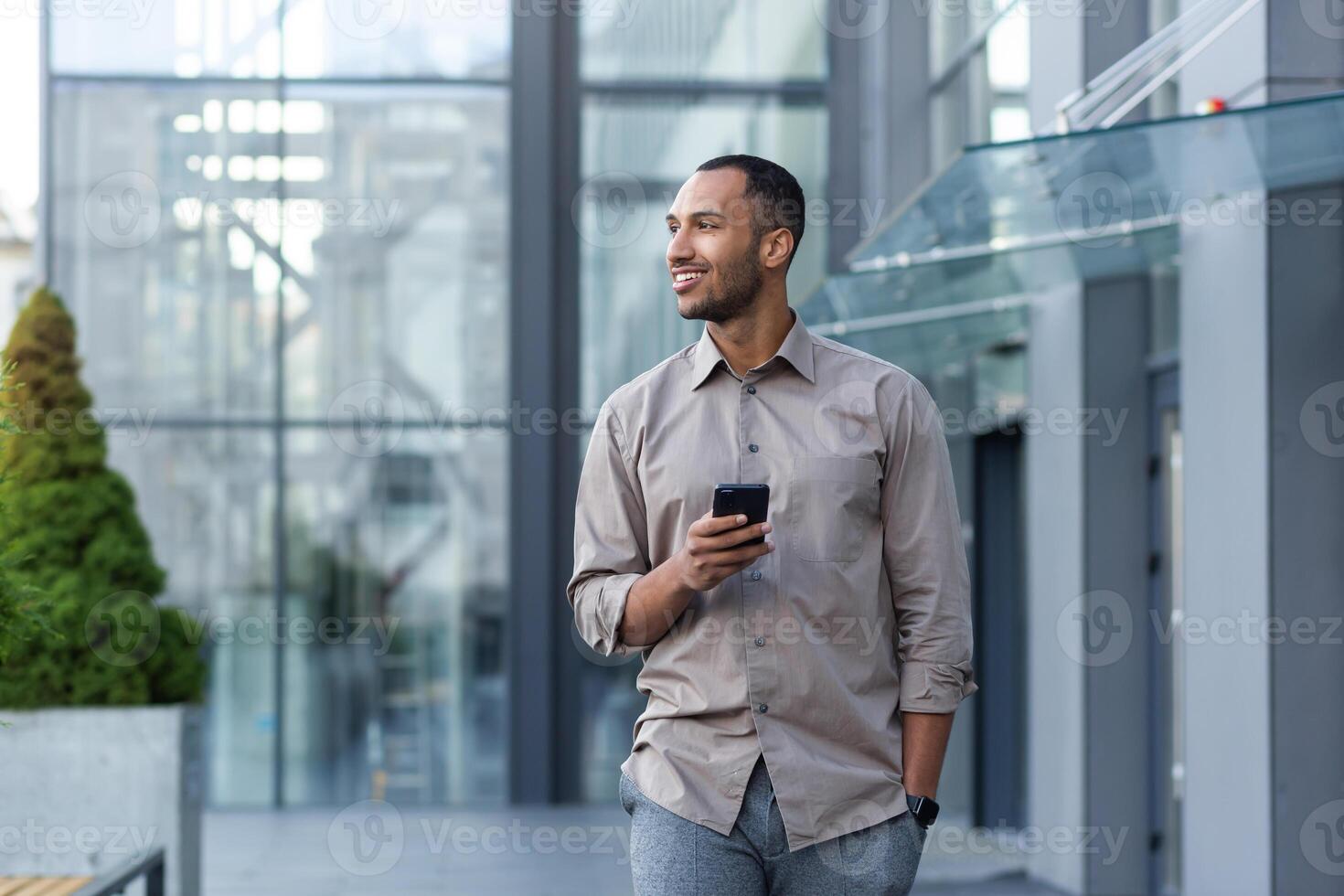 sonriente Hispano hombre caminando abajo calle cerca moderno oficina edificio, persona de libre dedicación empresario mirando lejos participación móvil teléfono foto