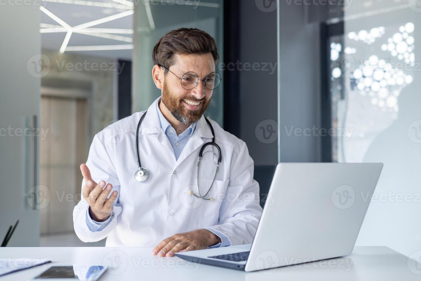 A smiling young male doctor sits in the office of the clinic at a desk, communicates and consults online through a laptop. photo