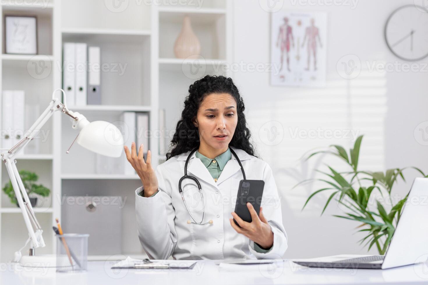 A female doctor in a white lab coat shows a puzzled expression while looking at her smartphone in a modern clinic office. Plants and anatomy charts in the background. photo