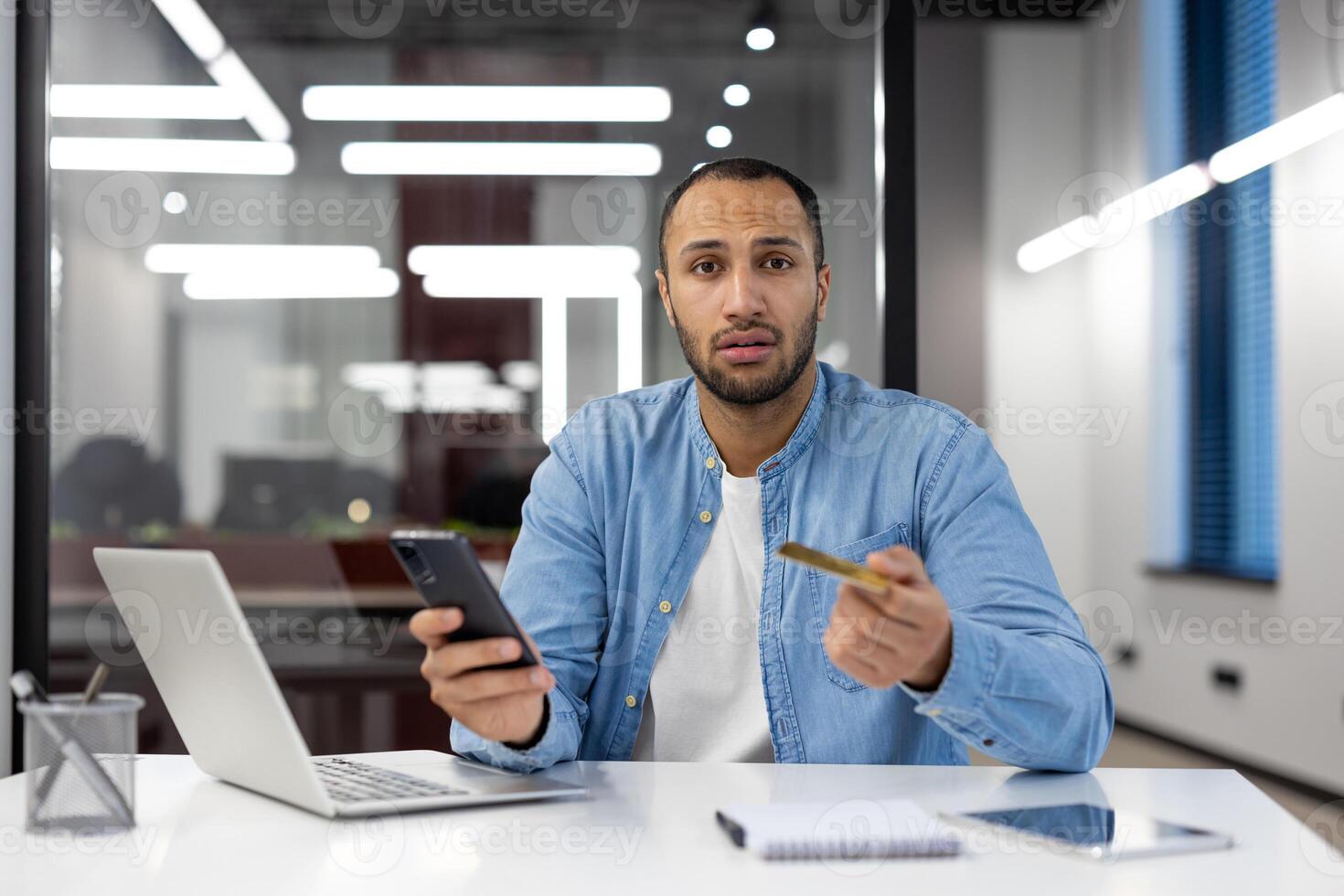 un hombre es sentado a un escritorio con un ordenador portátil y un célula teléfono. él es vistiendo un azul camisa y un blanco camisa. él es participación un crédito tarjeta en su mano. concepto de trabajo y productividad foto