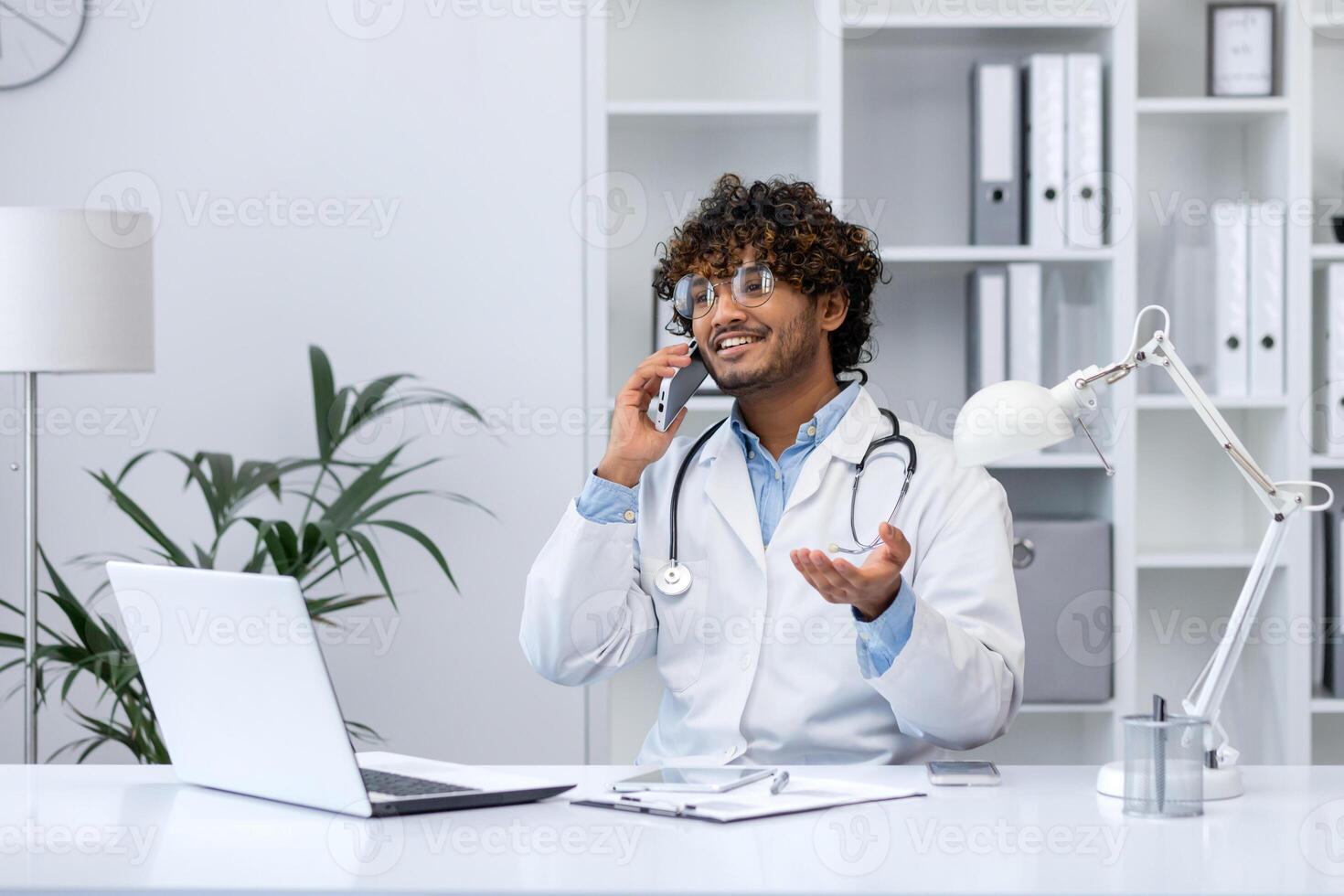 joven exitoso Hispano médico dentro el clínica en el médico oficina alegremente comunica hablando en el teléfono, el clínica trabajador se sienta a el escritorio trabajando con un computadora portátil, consulta pacientes foto