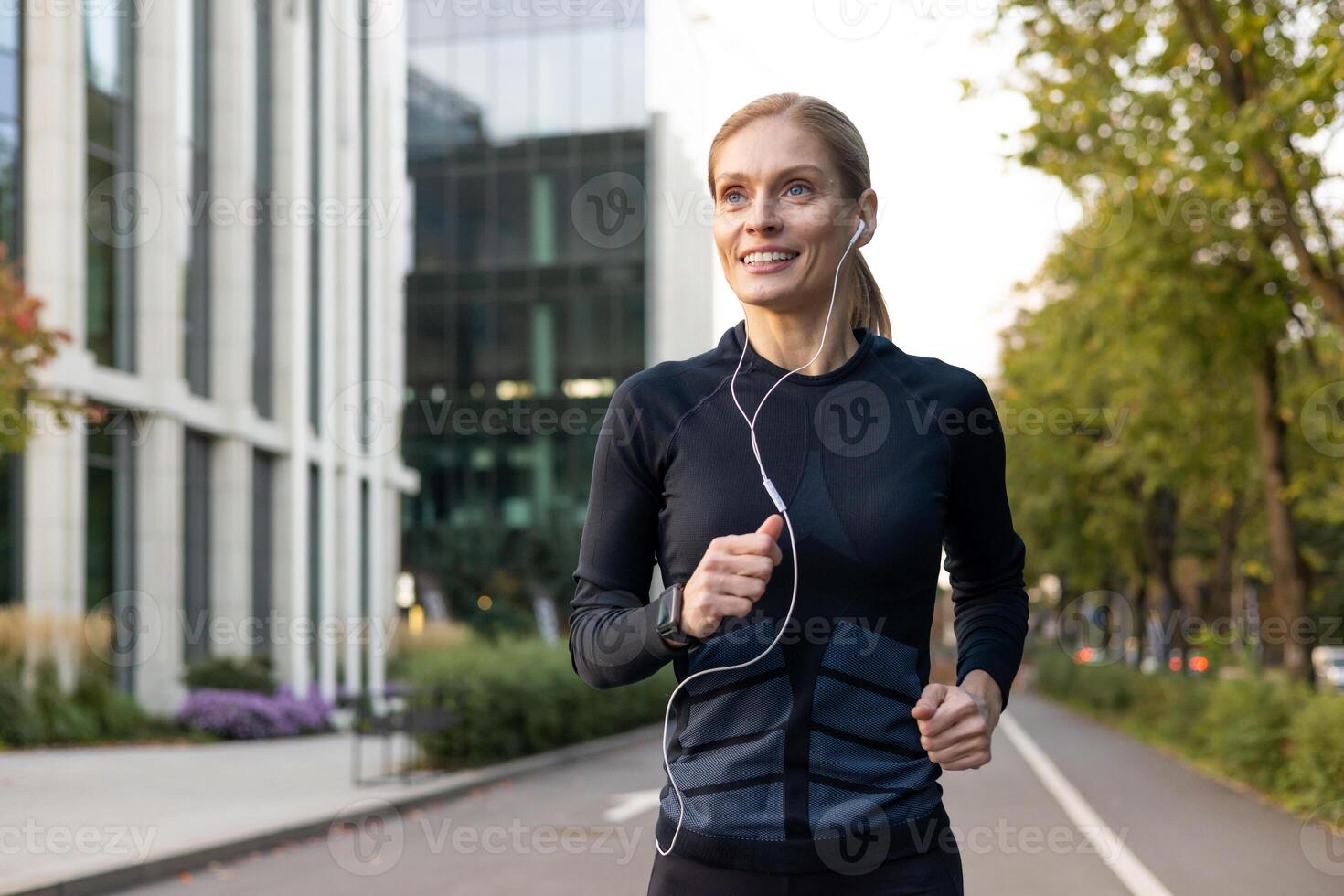 maduro experimentado adulto mujer corriendo en auriculares en chandal fuera de moderno edificio cerca árboles, contento con rutina de ejercicio deportista sonriente activo estilo de vida. foto