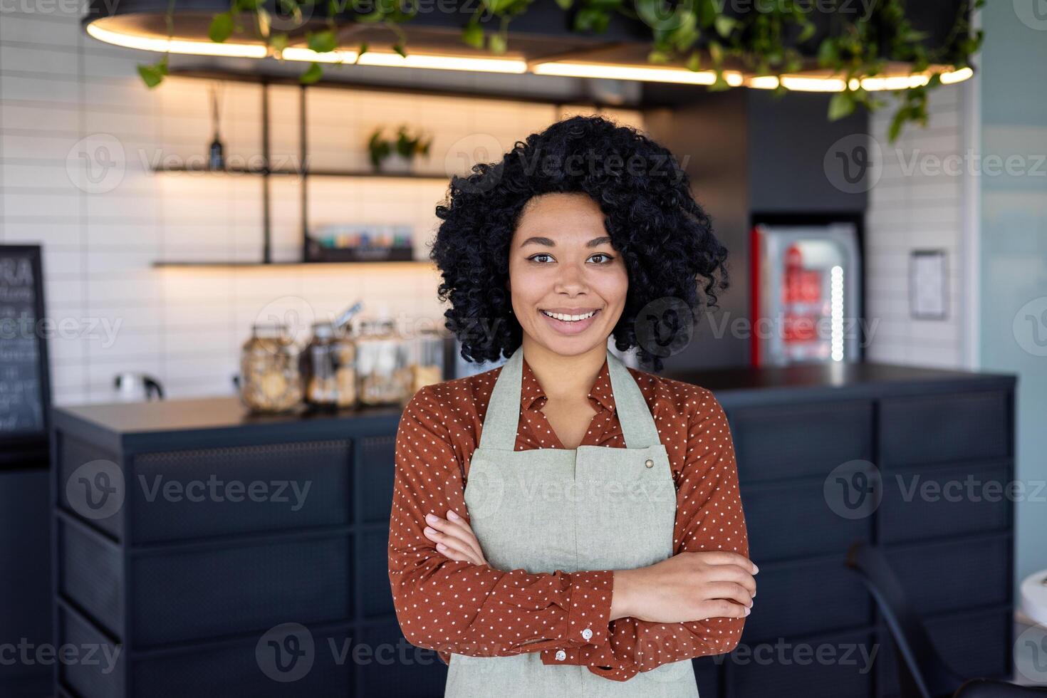 Satisfied black woman in apron standing with crossed arms on chest on background of bar counter. Confident small business owner in service sector ready for accepting clients in beginning of day. photo