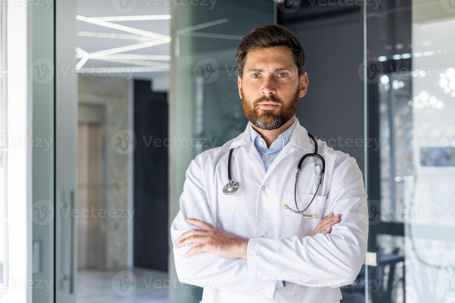 Portrait of a self-confident male doctor standing in a hospital room in a white coat, crossing his arms on his chest and seriously looking at the camera. photo
