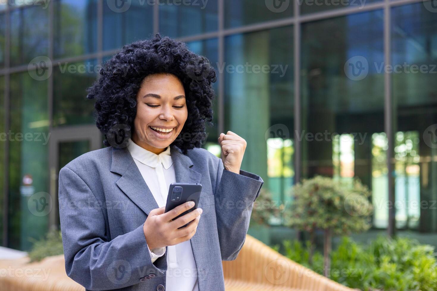 Close-up photo of young African American woman in suit standing near office center, holding and looking at mobile phone, happy showing victory gesture with hand.