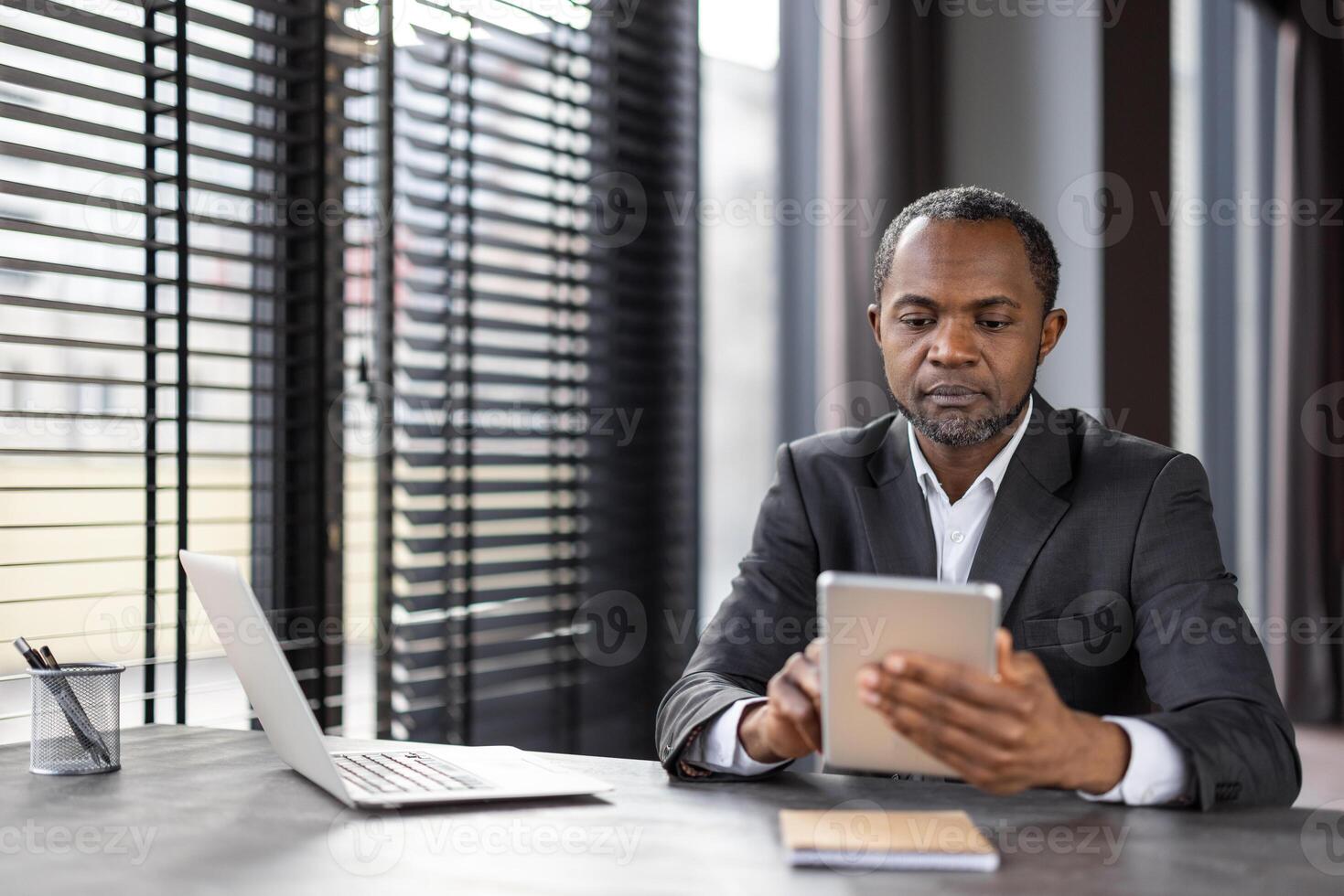 Focused entrepreneur scrolling internet through digital tablet while working by window with jalousie. Attentive financial manager entering data about company bills in corporate system application. photo