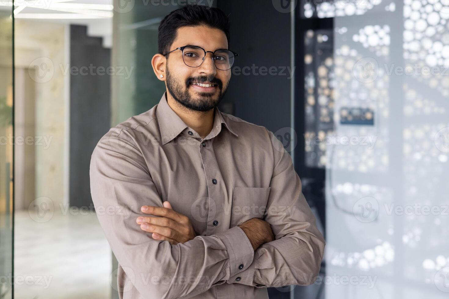 A professional young man with a beard and glasses, smiling confidently in a well-lit, contemporary office setting. photo