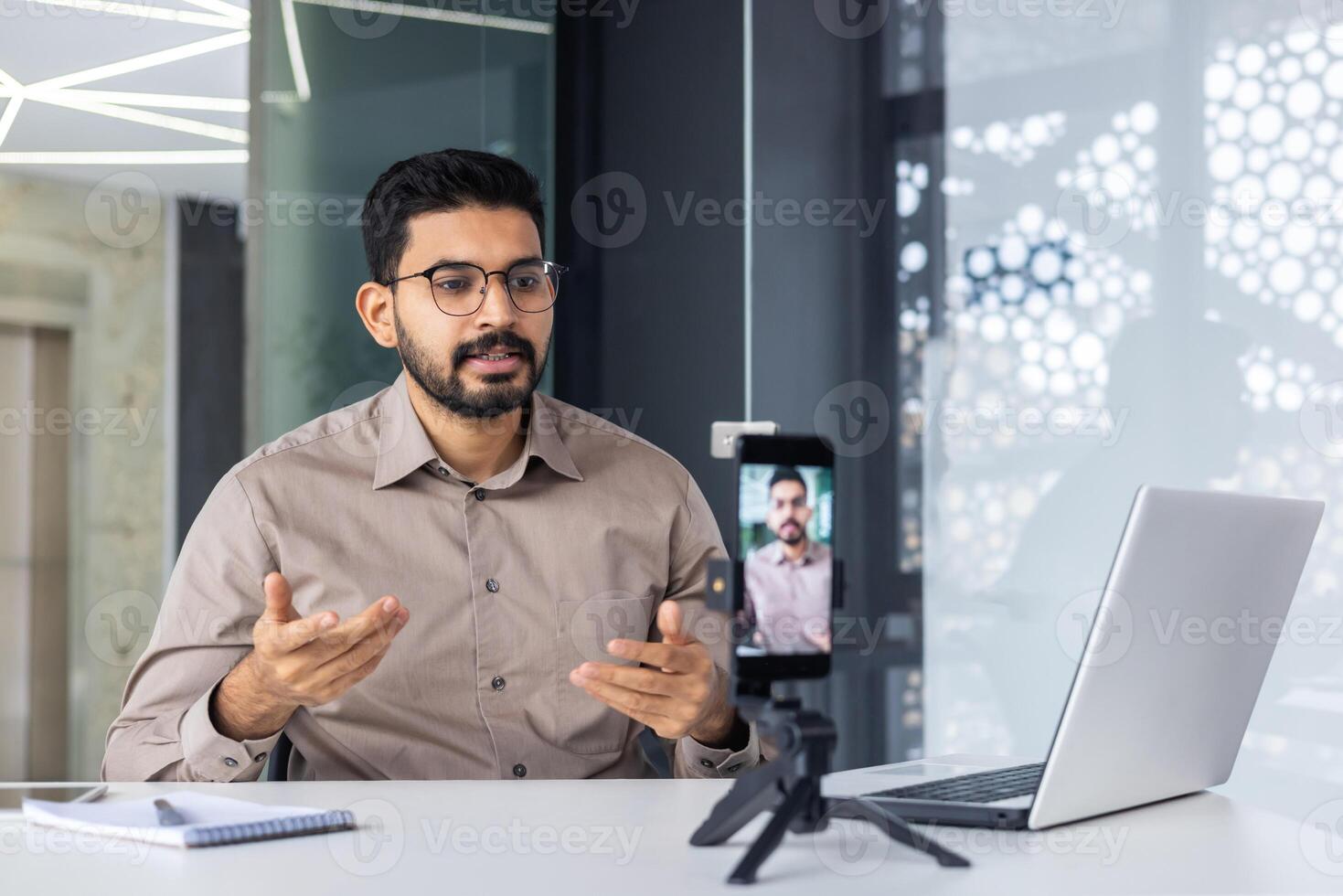 Successful business trainer teacher recording online training course, man inside office at workplace using phone and tripod, sitting at desk with laptop. photo