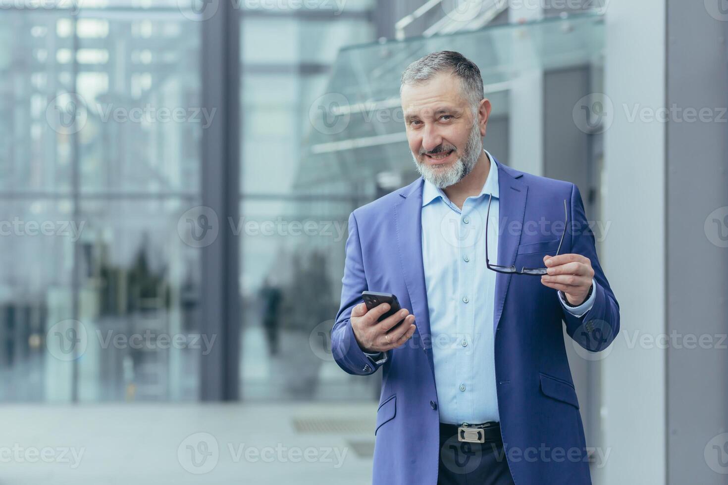 Portrait senior handsome gray-haired male teacher, professor, lecturer stands in a suit in a modern campus, holds a phone and glasses. He looks at the camera, smiles. photo
