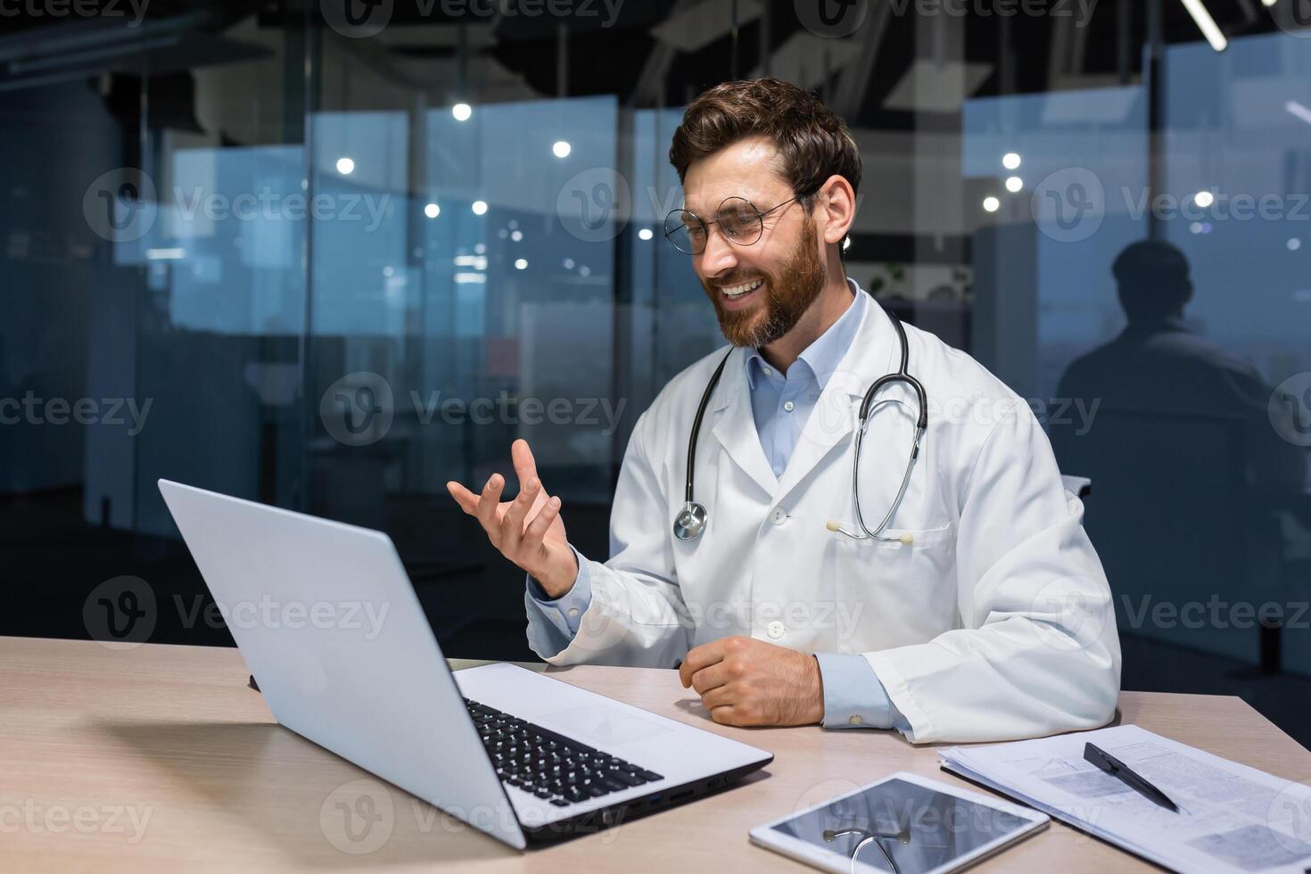 Experienced mature doctor working remotely, senior man in medical gown working online consulting patients inside modern clinic, doctor using laptop for online call. photo