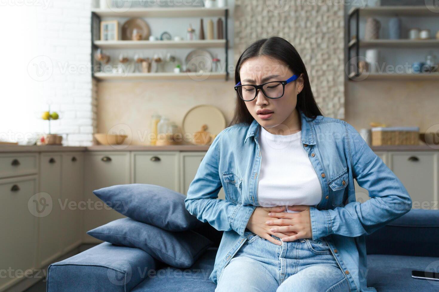 Stomach disorder. A young beautiful Asian woman holds her stomach with her hands, feels severe pain. Sitting at home on the sofa. photo