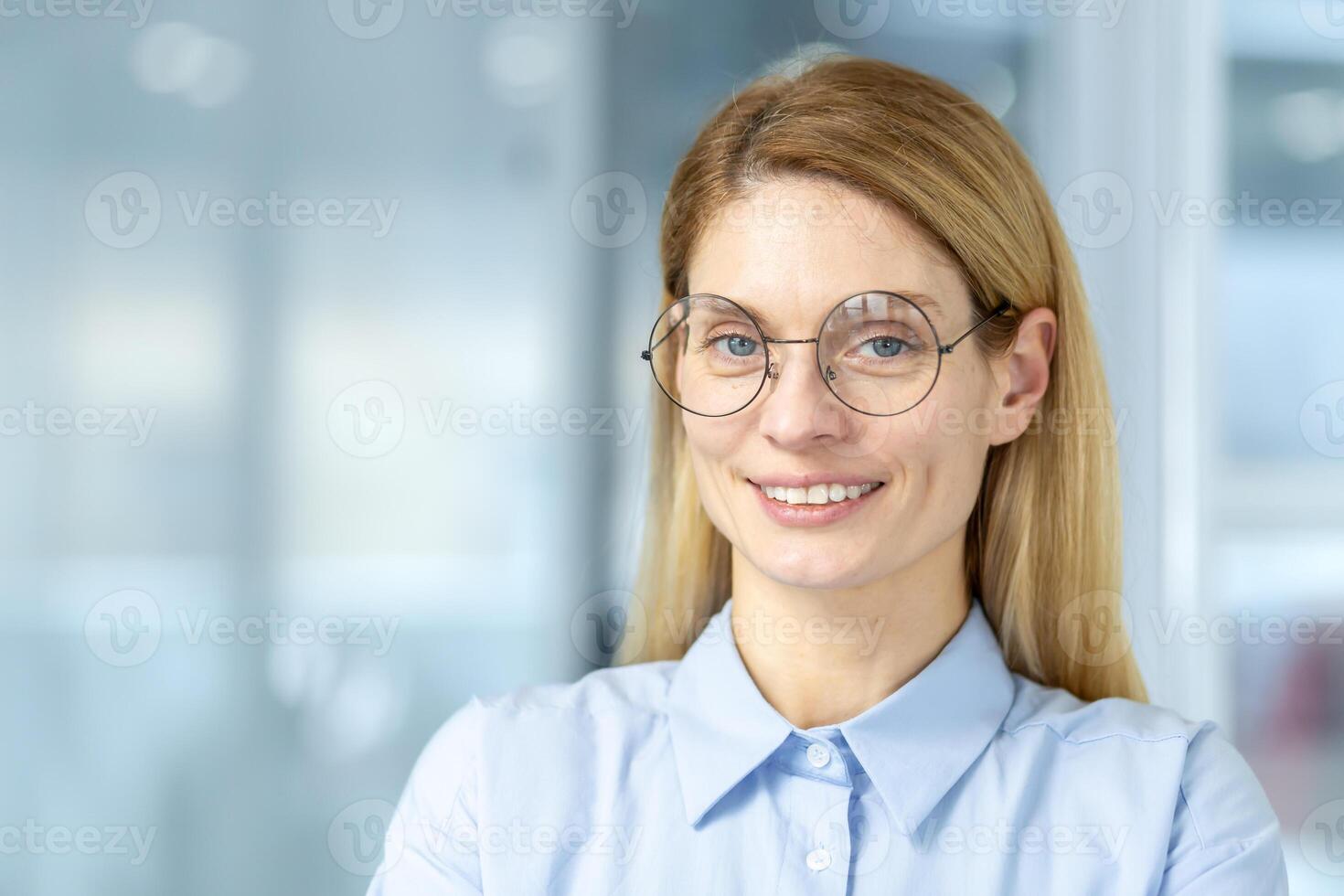 Portrait of blue eyed executive in eyewear looking at camera with wide smile on blurred background. Happy woman in formal clothes standing with satisfied facial expression in business office. photo