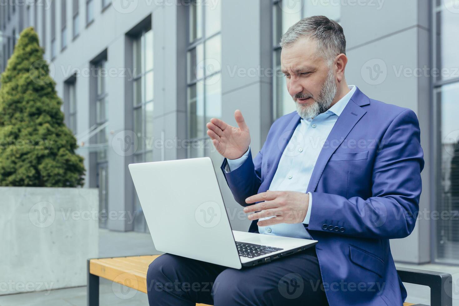 Successful senior male businessman conducts a conference from a laptop. Sitting outside in a suit on a bench near an office center, waves his hands, explains. photo