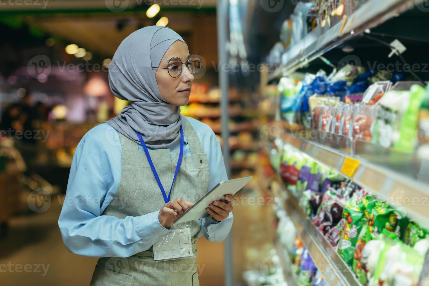 mujer vendedor en súper mercado en hijab con tableta comprobación productos utilizando bolsillo computadora, musulmán mujer cerca estantería con productos y bienes foto