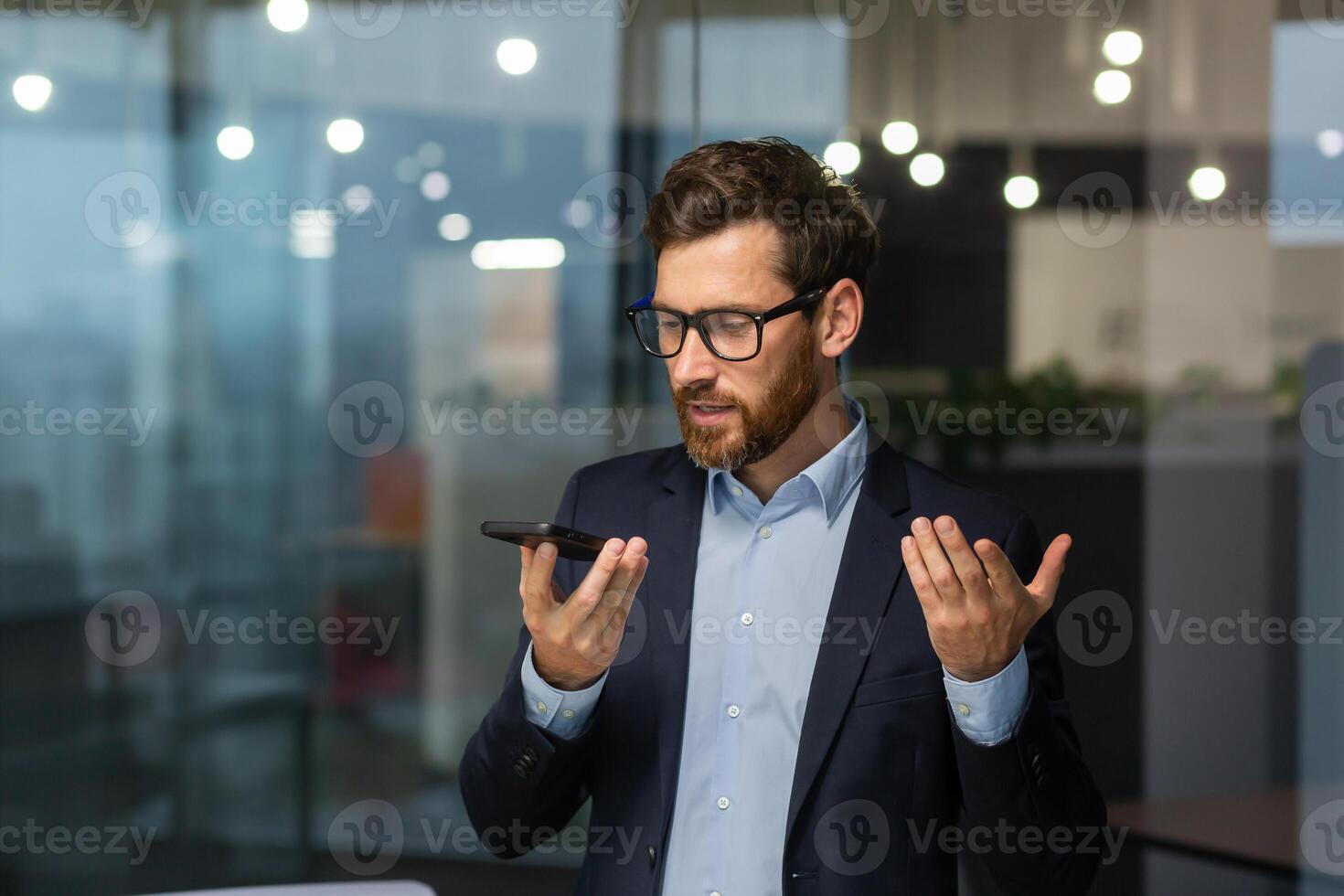 Friendly and smiling businessman in office recording audio message, man using smartphone app for communication, investor in business suit near window inside building. photo
