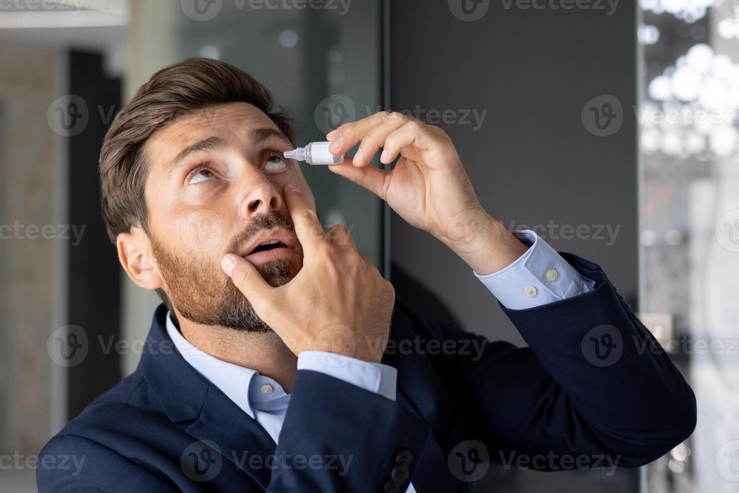 Close-up photo of a young businessman sitting in the office at a desk in a suit and instilling medicine into his eye, treating inflammation, pain, chronic fatigue.