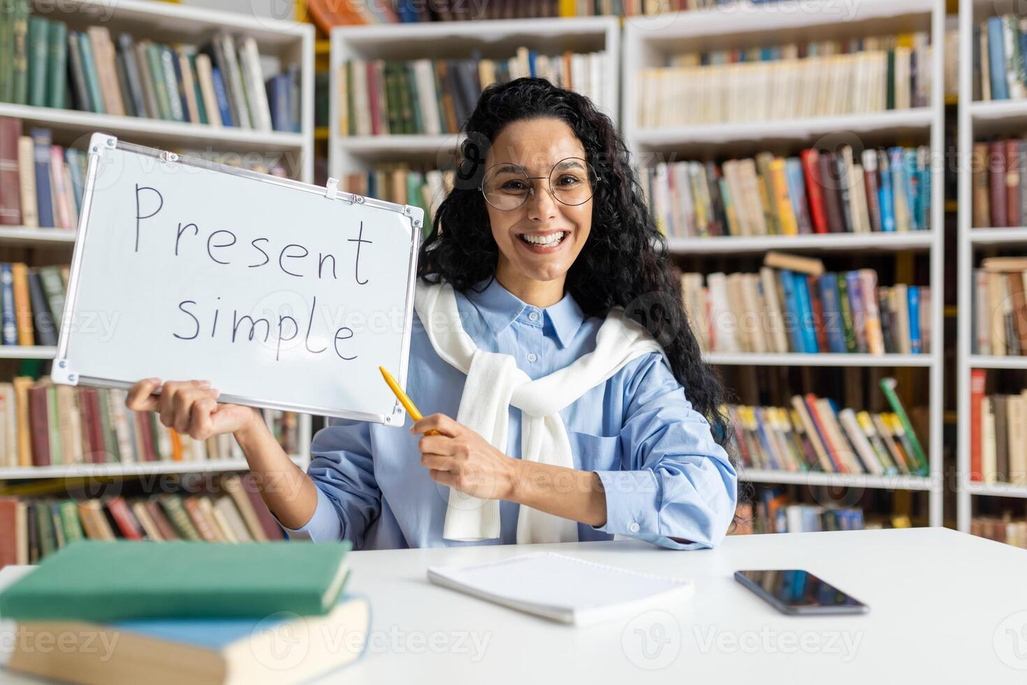 Cheerful educator teaching the present simple tense, using a whiteboard in front of a bookshelf-filled library. photo