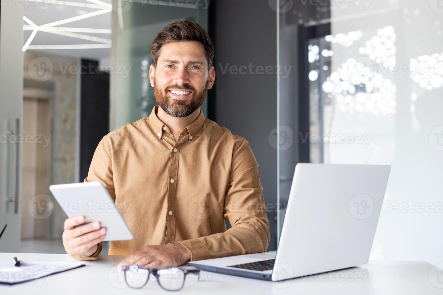 Portrait of a young male programmer working in the office at a laptop and holding a tablet in his hands, smiling and confidently looking at the camera. photo