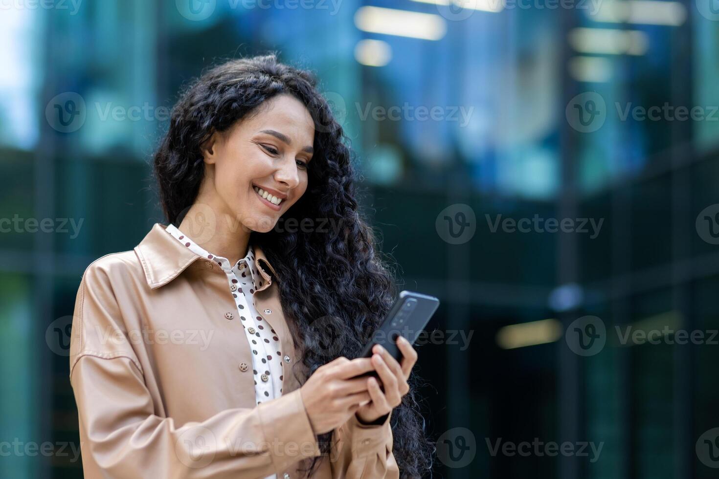 Young beautiful hispanic woman walking in the city, business woman holding phone in hands using smartphone app, woman smiling contentedly and happy outside office building with curly hair photo