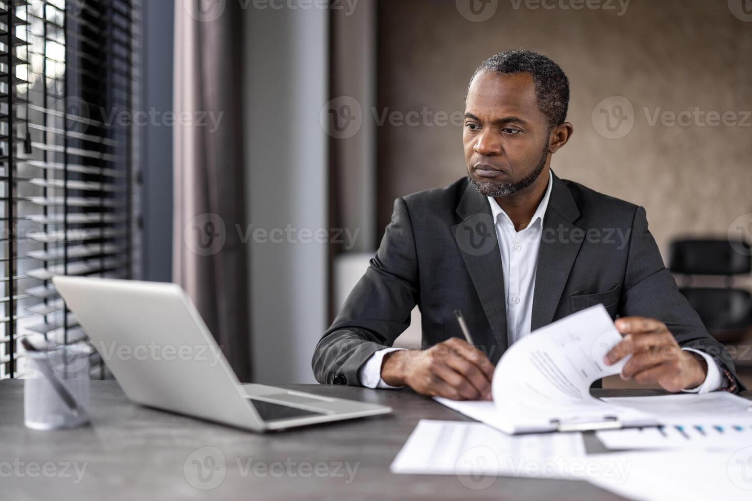 Focused professional analyzing and working on reports with laptop and notepad in a modern office setting. photo