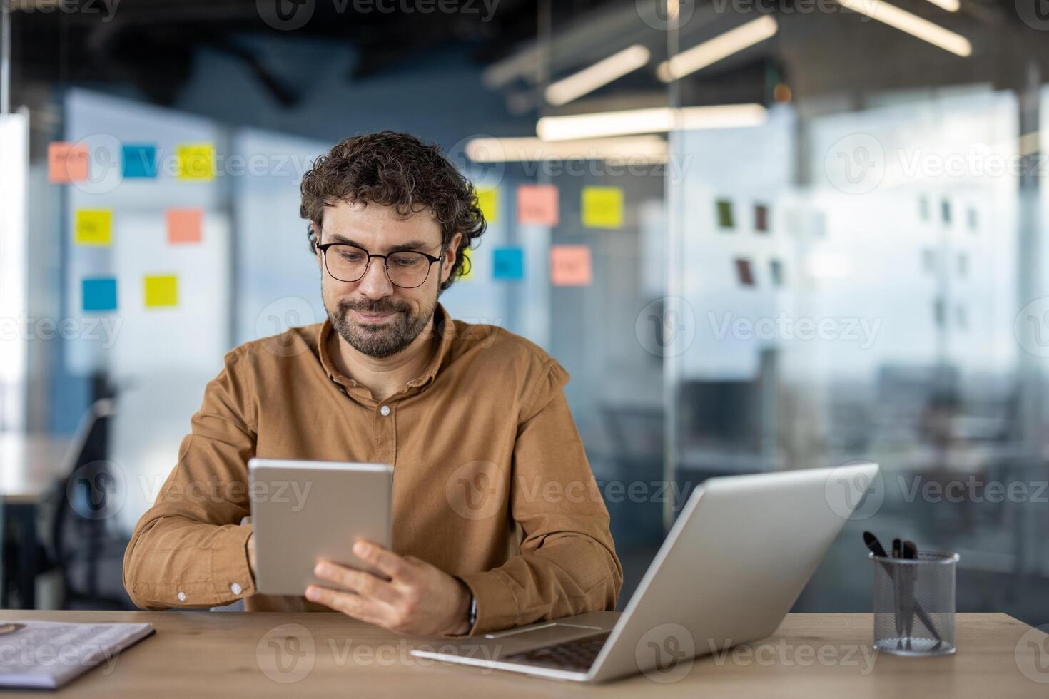 Focused male business professional using a tablet and laptop at a modern workspace with sticky notes in the background. photo