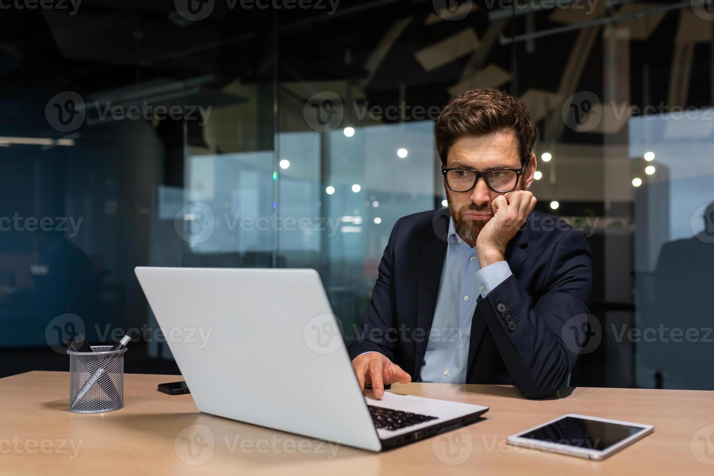 sad and depressed businessman in depression working inside modern office building, man in business suit with glasses and beard using laptop for work, boss disappointed with work result. photo