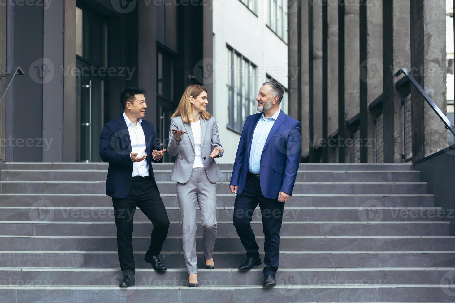 Interracial business meeting. Senior gray-haired man, young Asian man and one businesswoman in business suits walking up the stairs of a modern office center, talking to each other, discussing a deal photo