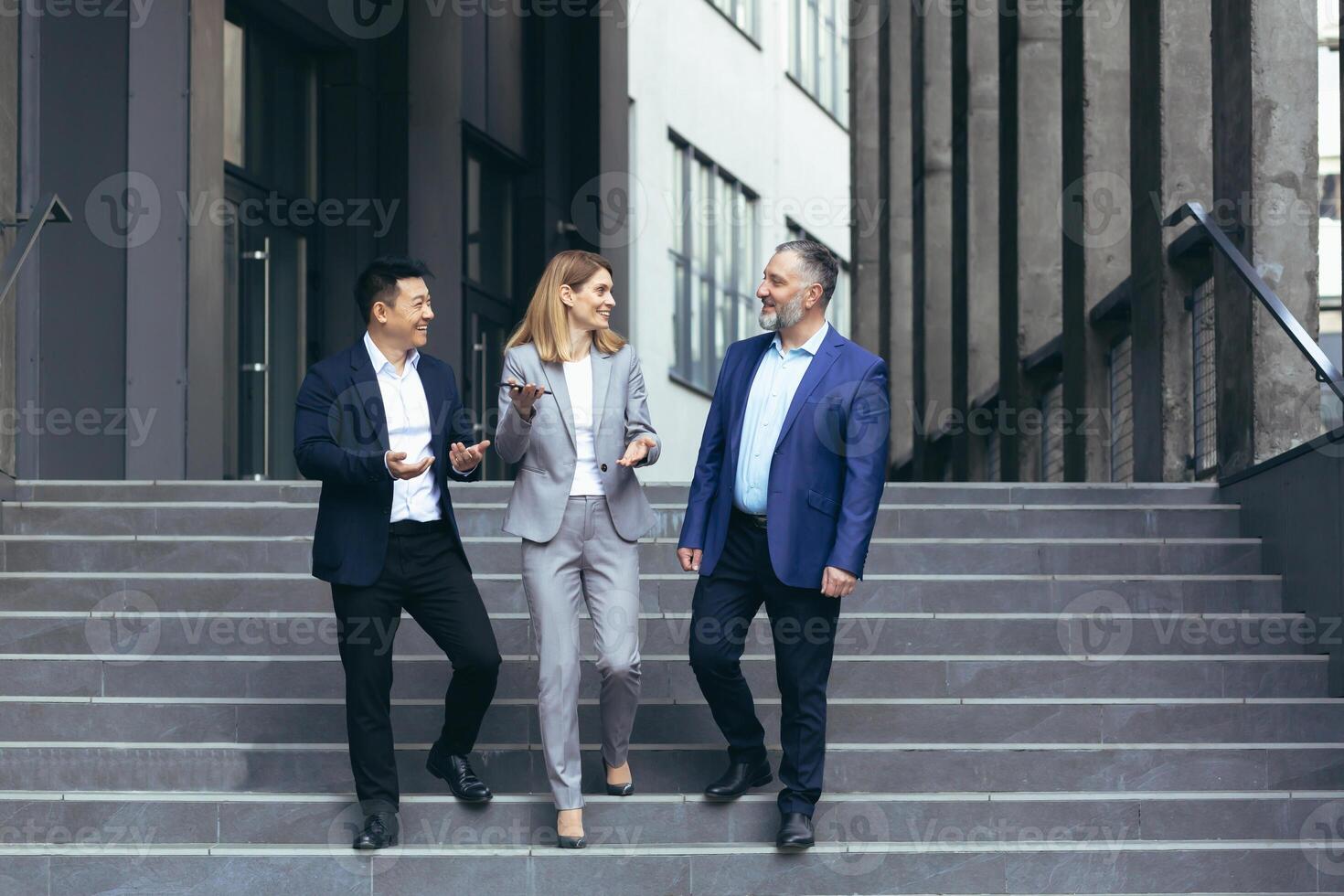 interracial negocio reunión. mayor canoso hombre, joven asiático hombre y uno mujer de negocios en negocio trajes caminando arriba el escalera de un moderno oficina centro, hablando a cada otro, que se discute un acuerdo foto