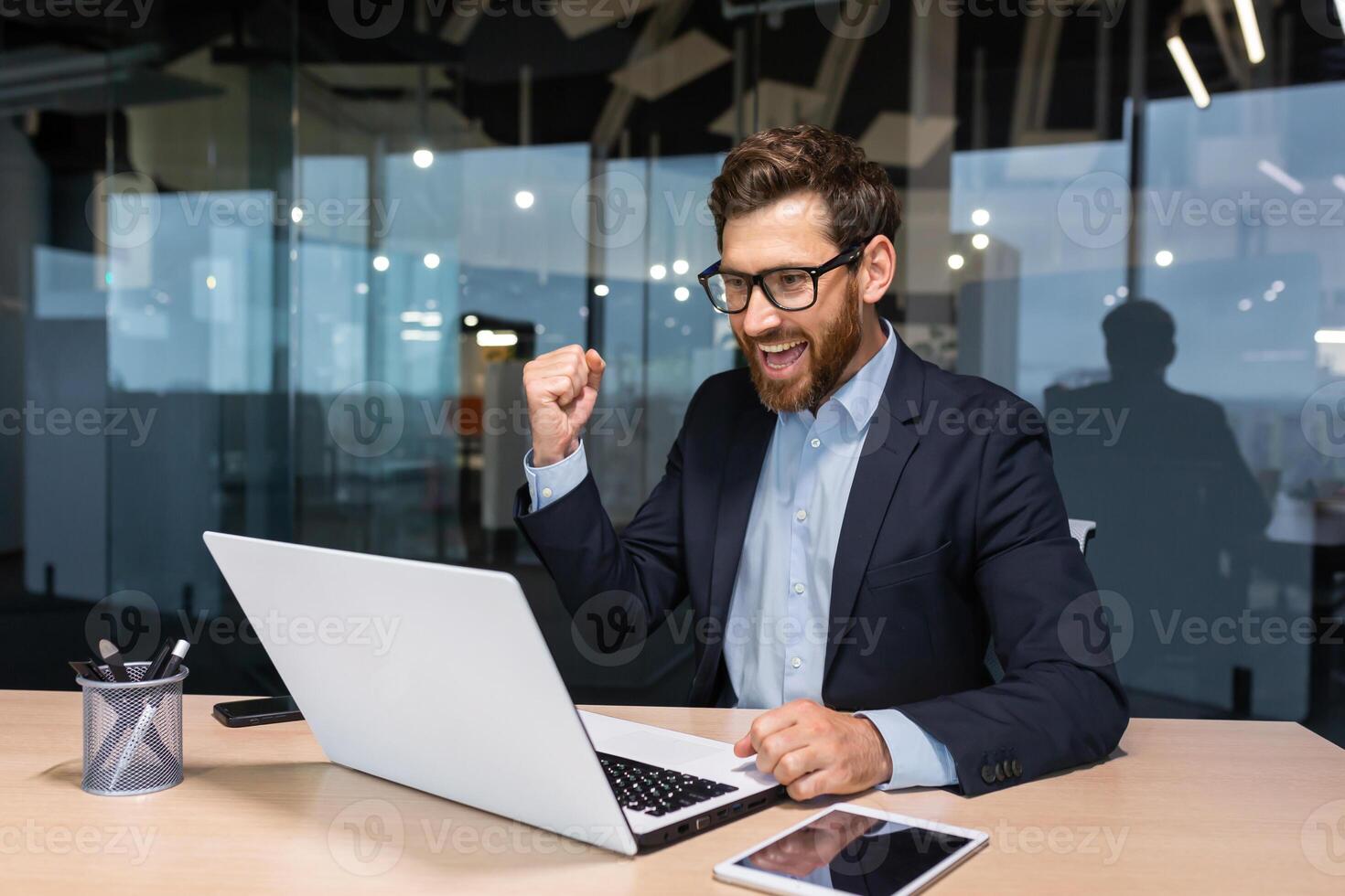 Successful businessman celebrating victory and good business achievement, boss using laptop reading notification from screen and holding hand up smiling triumph gesture. photo