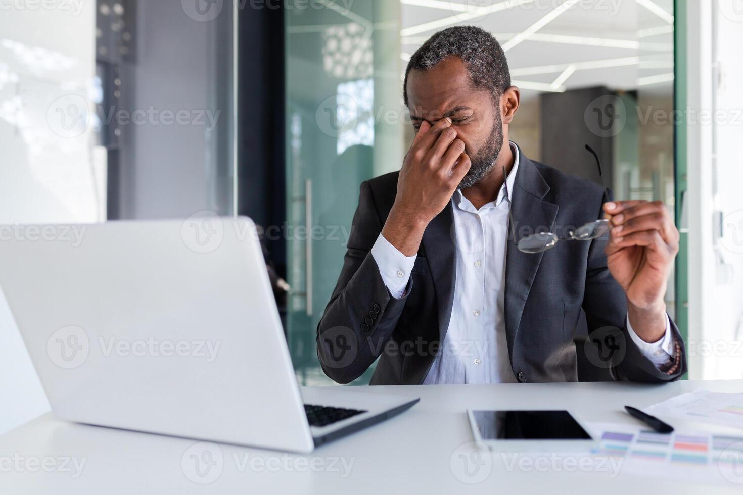 sobrecargado de trabajo hombre a lugar de trabajo dentro oficina, africano americano empresario trabajando sentado con ordenador portátil a trabajar, jefe tomando apagado lentes y masajear ojos. foto