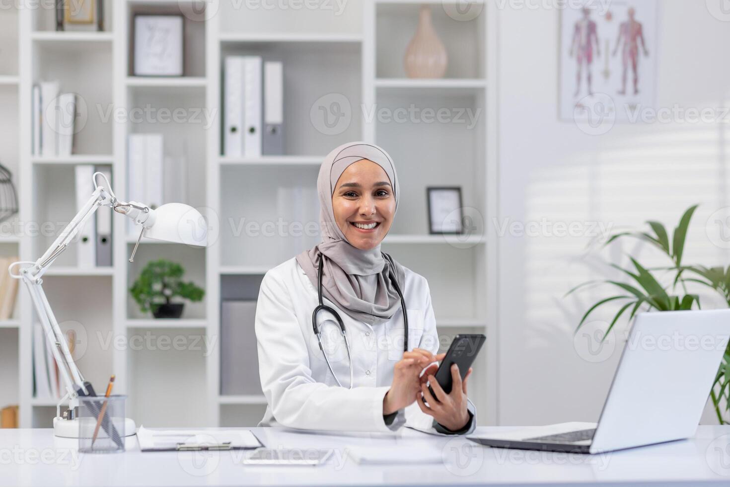 A cheerful female healthcare professional in a hijab sits at her office desk, using a smartphone and looking at a laptop, conveying a sense of accessibility and modern healthcare. photo