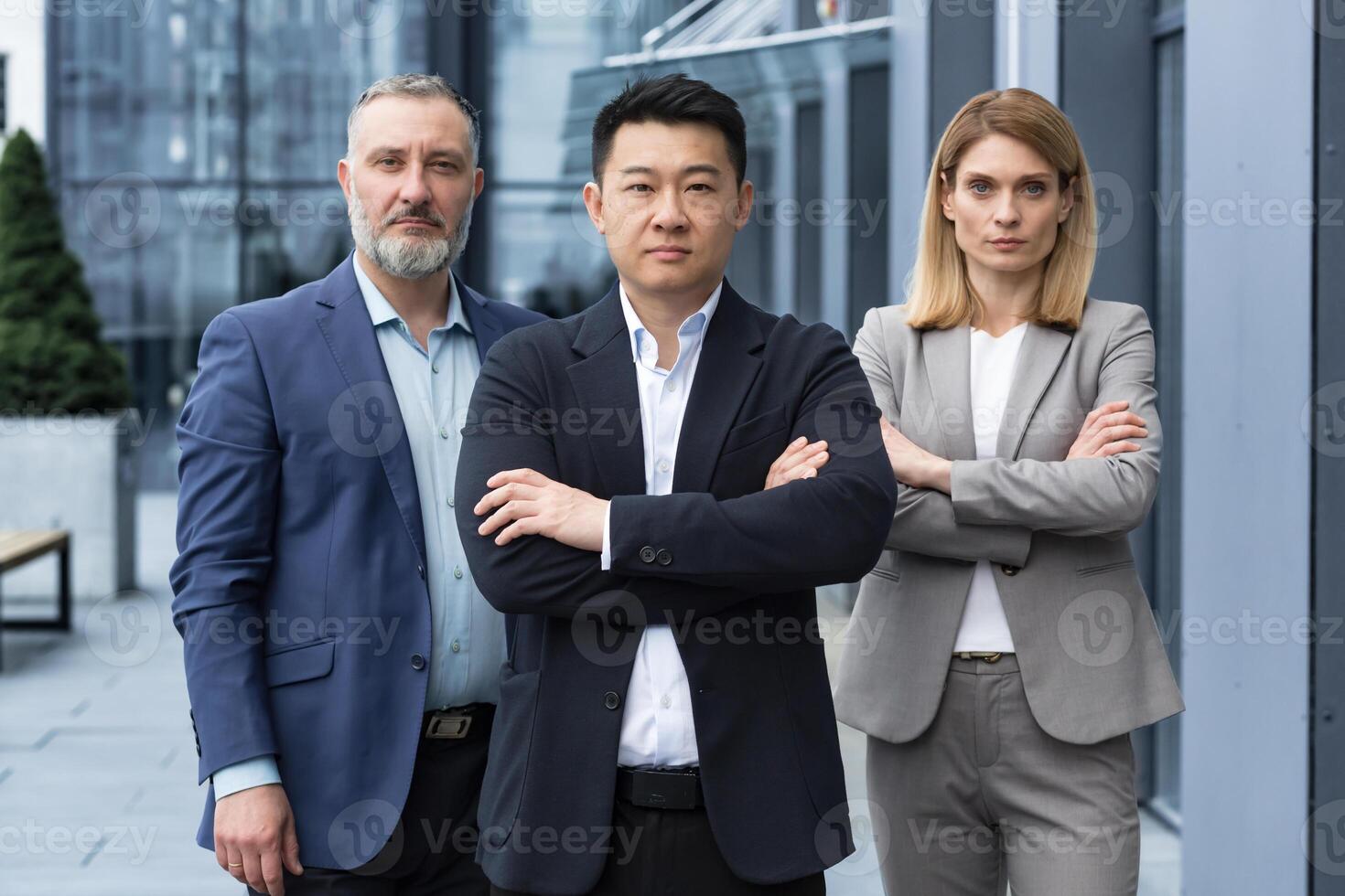 Successful and serious diverse team of three business people, man and woman focused looking at camera with arms crossed, portrait of co-workers outside office building photo