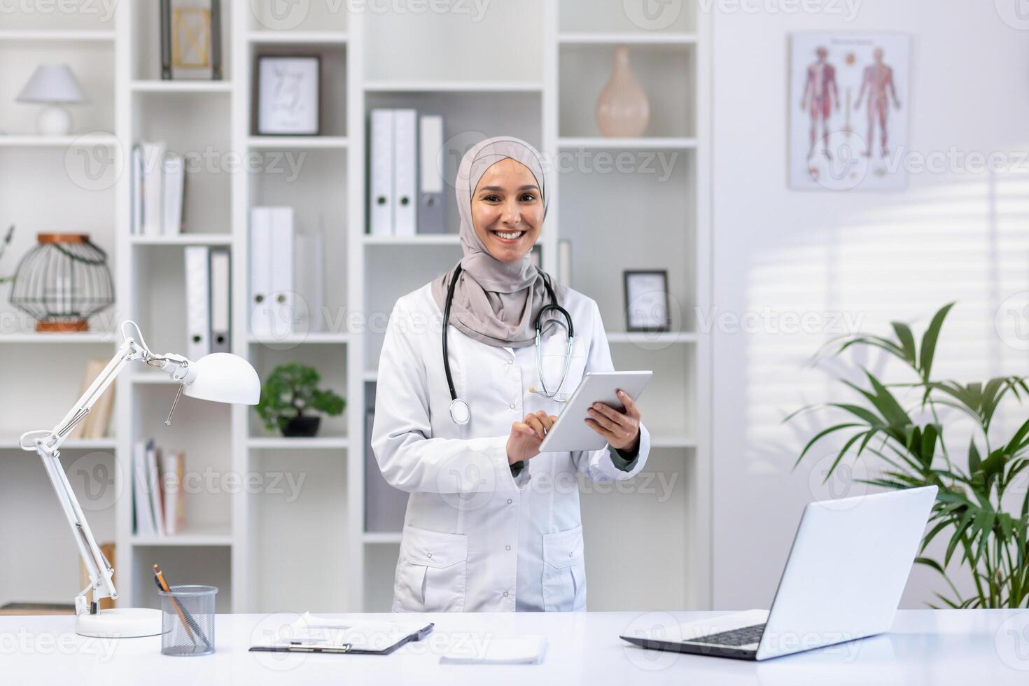 sonriente hembra musulmán médico en un hijab y blanco Saco utilizando un tableta en un brillante médico oficina, retratar cuidado de la salud profesionalismo. foto