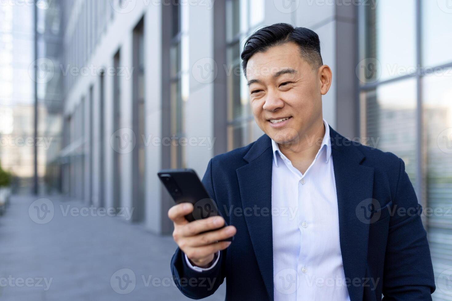 Asian smiling young man walking down the street near an office building and using a mobile phone. photo