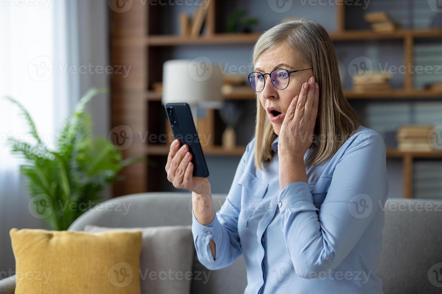 Mature woman with glasses feeling surprised holding a phone, displaying a shocked expression in a cozy living room setting. photo